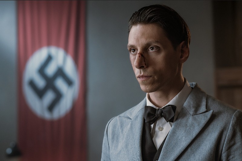 Mężczyzna w jasnym garniturze i muszce stoi w pomieszczeniu z widoczną raną na nosie. Na rozmytym tle wisi duża flaga nazistowska ze swastyką.