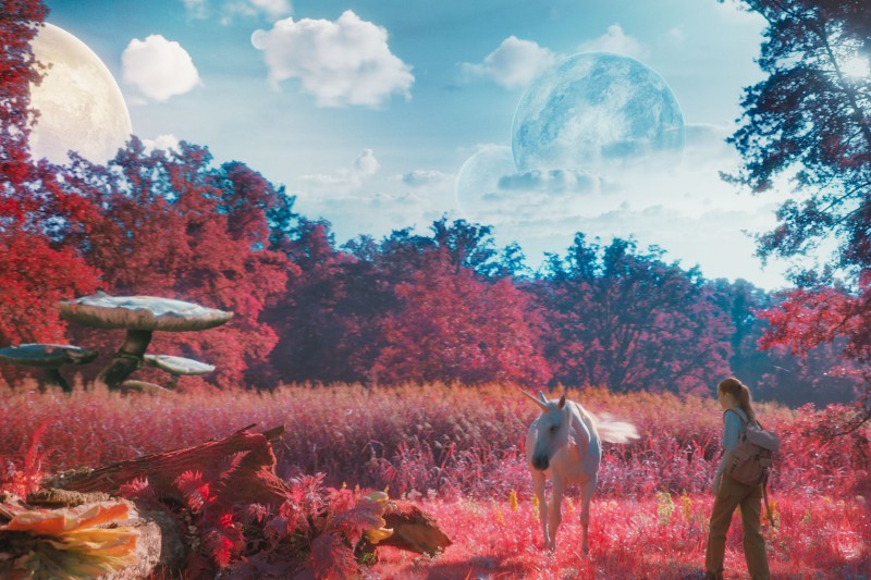 Młoda kobieta stoi w surrealistycznym różowym lesie z gigantycznymi grzybami, naprzeciwko białego jednorożca. Niebieskie planety i chmury wypełniają niebo, tworząc marzycielski, fantastyczny krajobraz.