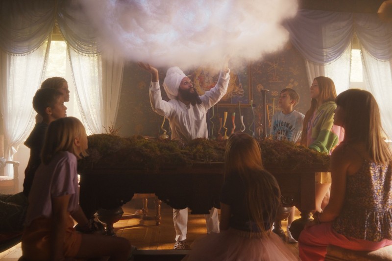 Osoba w czapce kucharskiej dramatycznie gestykuluje pod świecącą chmurą nad stołem, otoczona dziećmi siedzącymi i obserwującymi potrawę w nasłonecznionym, fantazyjnym pokoju z drapowanymi zasłonami.