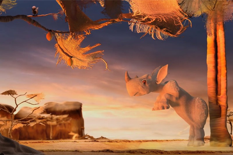 Stylizowany, animowany mały nosorożec stoi na tylnych nogach w pobliżu palm o zachodzie słońca, a inne małe zwierzę siedzi na drzewie powyżej. Wszystko to dzieje się w surrealistycznym, pustynnym krajobrazie.
