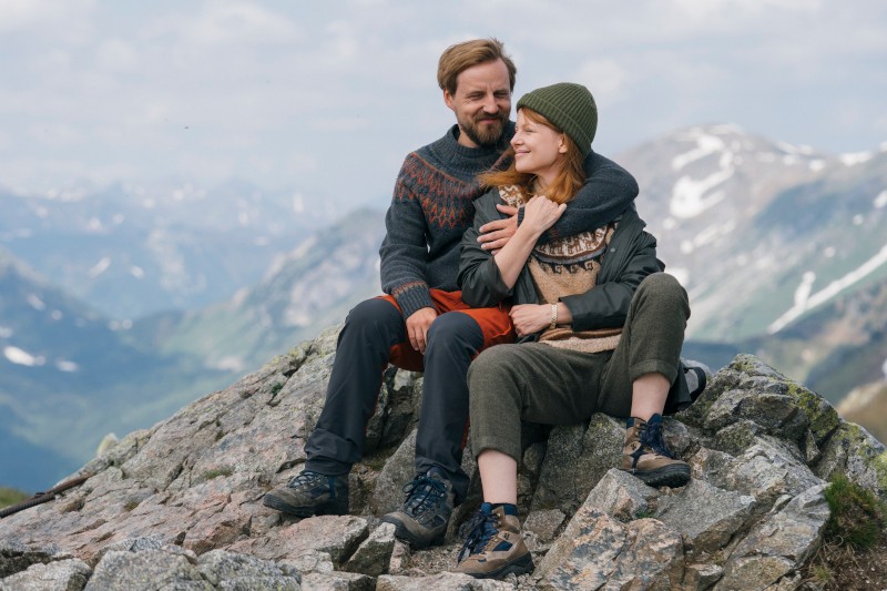 Para w ciepłych ubraniach siedzi na skalistym terenie na szczycie góry, obejmując się i uśmiechając. W tle widać ośnieżone szczyty i zachmurzone niebo.