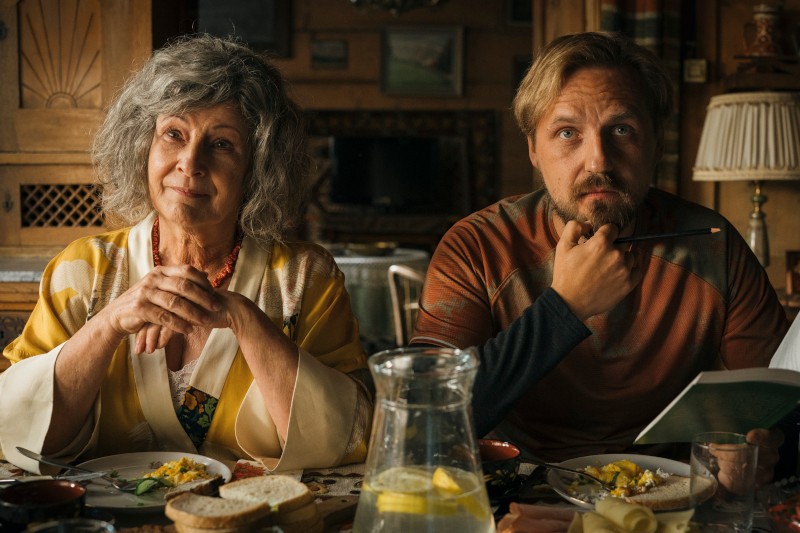 Starsza kobieta i młodszy mężczyzna siedzą przy stole jadalnym z talerzami jedzenia, chlebem i dzbankiem wody z cytryną. Kobieta wygląda na zadowoloną, podczas gdy mężczyzna trzyma ołówek i notatnik, sprawiając wrażenie zamyślonego.