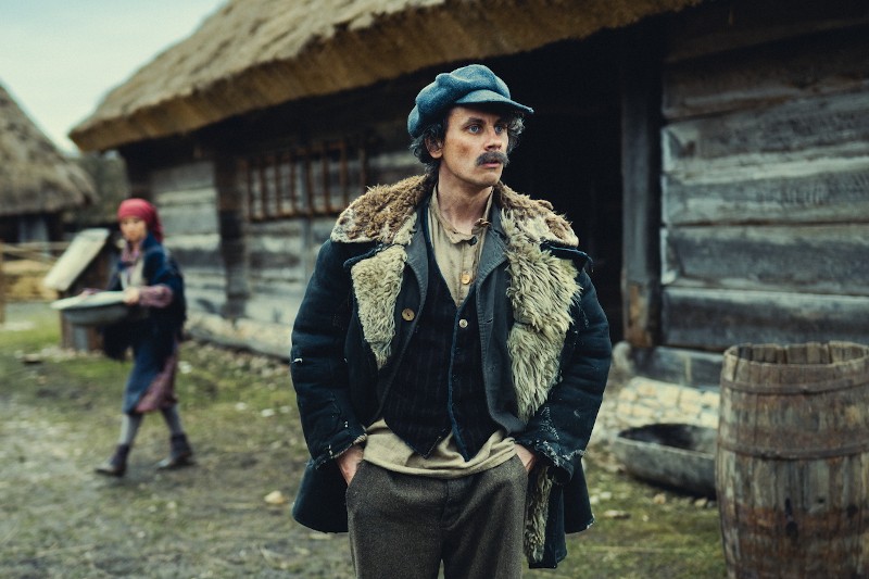 Mężczyzna w staromodnym, warstwowym ubraniu i niebieskiej czapce stoi przed rustykalnymi drewnianymi budynkami ze strzechą. Ma poważny wyraz twarzy. W tle osoba w czerwonej chuście na głowie niesie tacę.