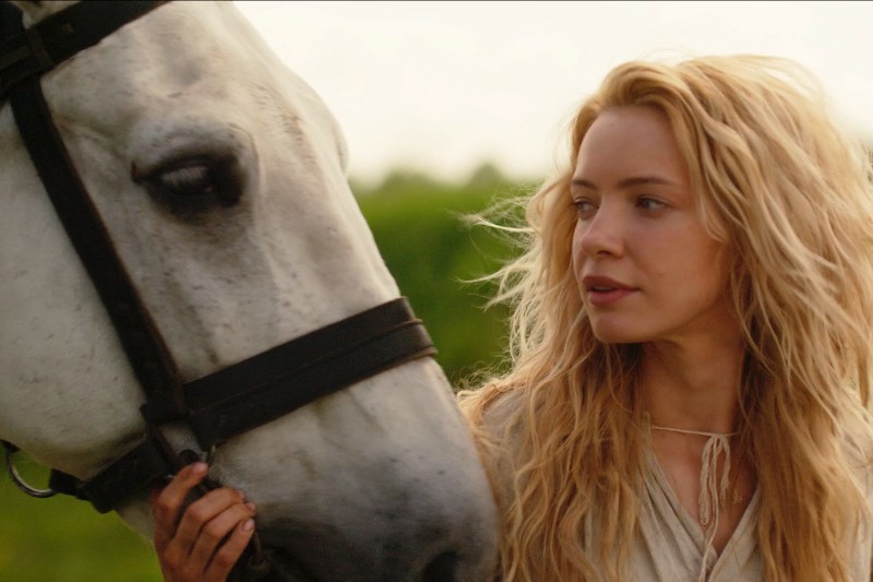 Kobieta o długich, falujących blond włosach stoi blisko białego konia, delikatnie trzyma jego uzdę i patrzy na konia spokojnym wzrokiem; w tle rozmazują się zielone liście.