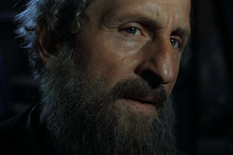 Zbliżenie mężczyzny z gęstą, siwiejącą brodą i kręconymi włosami, patrzącego lekko poza kamerę. Jego wyraz twarzy jest poważny, a oświetlenie podkreśla fakturę jego skóry i zarostu.