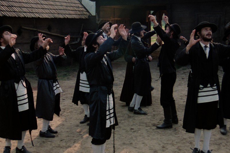 Grupa mężczyzn ubranych w tradycyjne stroje żydowskie stoi na zewnątrz na ziemi, podnosząc ramiona w jednolitym geście, sprawiając wrażenie, jakby wspólnie się modlili lub tańczyli.