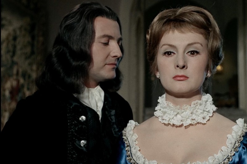 Mężczyzna o długich, ciemnych włosach i w czarnym płaszczu stoi tuż za kobietą o krótkich, jasnych włosach, ubraną w biały kołnierzyk z falbaną i niebieską sukienkę. Oboje są ubrani w stroje historyczne.