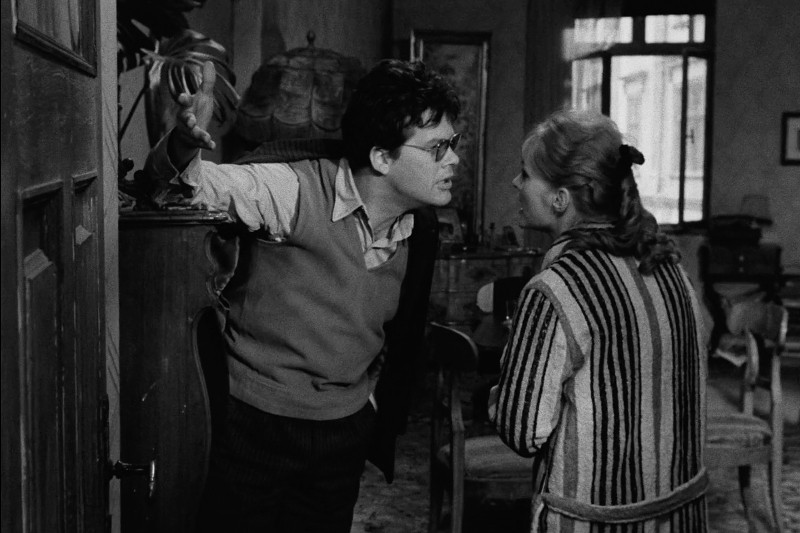 Mężczyzna w okularach pochyla się do przodu i gestykuluje, jednocześnie intensywnie rozmawiając z kobietą w pasiastej szacie w zagraconym, słabo oświetlonym pokoju o staromodnym wystroju.