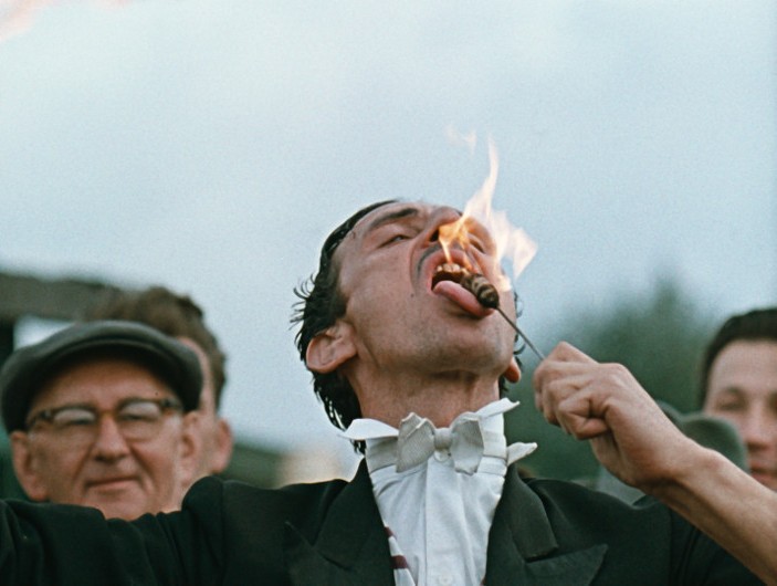 Mężczyzna ubrany w garnitur i muszkę wykonuje akt połykania ognia, trzymając płonący przedmiot przy otwartych ustach. Widzowie obserwują go w tle.