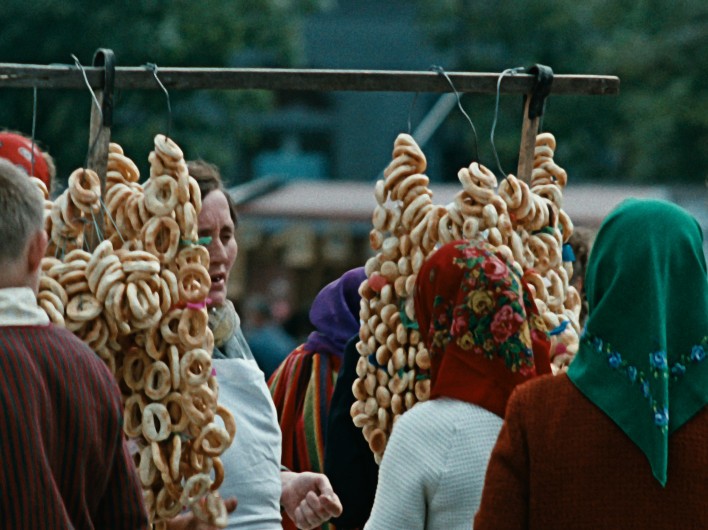 Kobiety w kolorowych chustach na głowach stoją wokół drewnianej ramy, na której eksponowane są sznury okrągłych bajgli lub krążków chlebowych na targu na świeżym powietrzu. W tle widać drzewa i inne stragany targowe.