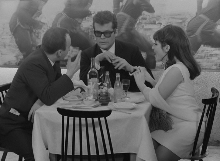 Trzy osoby siedzą przy stole zastawionym butelkami, szklankami i talerzami, pogrążone w ożywionej rozmowie. Jeden mężczyzna nosi okulary przeciwsłoneczne i garnitur, a kobieta w białej sukience gestykuluje ożywiona. Czarno-białe zdjęcie.