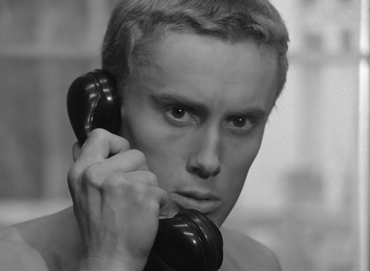 Osoba o krótkich jasnych włosach trzyma czarną słuchawkę telefonu przy uchu, patrząc uważnie przed siebie z poważnym wyrazem twarzy. Obraz jest czarno-biały.