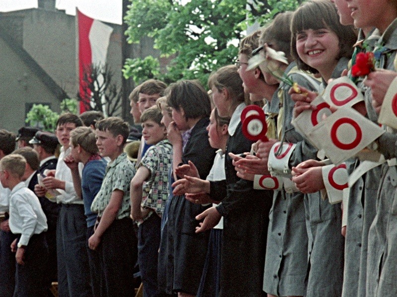 Grupa uśmiechniętych dzieci, niektóre trzymające okrągłe znaki i kwiaty, stoją w rzędzie na zewnątrz podczas wydarzenia publicznego. W tle widać drzewa i budynek z dużą czerwono-białą flagą.