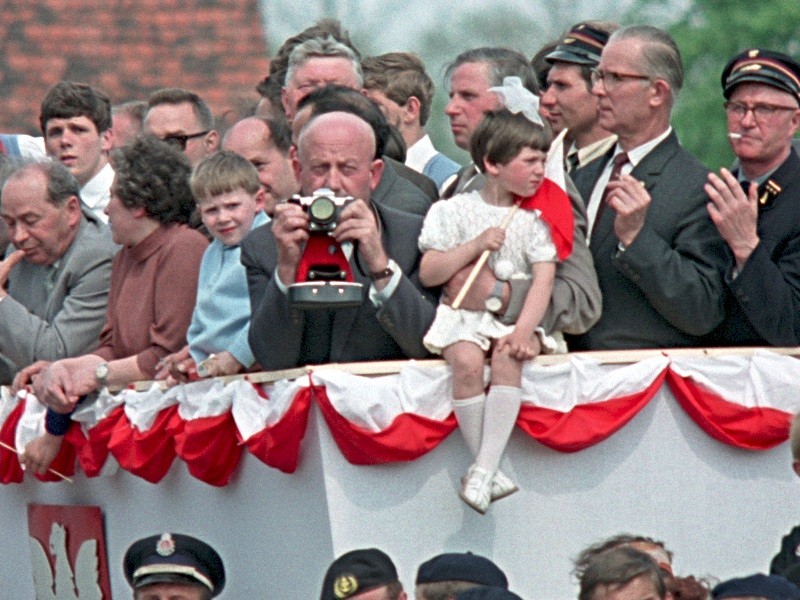 Tłum ludzi, w tym dorośli i dzieci, stoi za czerwono-biało udekorowaną barierą. Jeden mężczyzna robi zdjęcie, podczas gdy młoda dziewczyna w bieli siedzi na barierze trzymając zabawkę. Niektórzy mężczyźni noszą mundury i czapki.