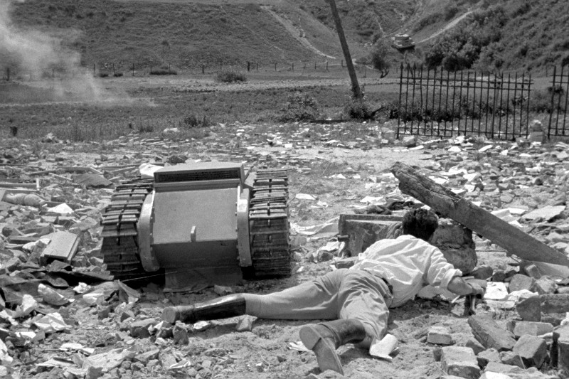 Osoba leży twarzą do dołu na skalistym podłożu obok małego pojazdu gąsienicowego na zewnątrz, na terenie zasypanym gruzem, z unoszącym się dymem i widocznymi wzgórzami w tle.