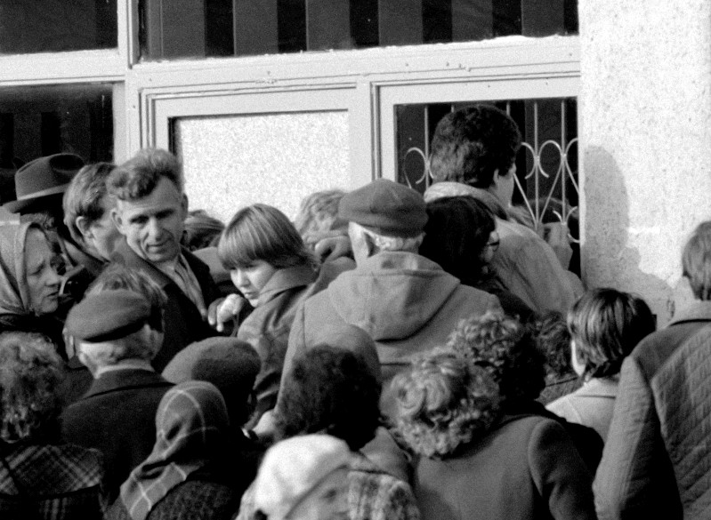 Czarno-białe zdjęcie przedstawia tłum ludzi zgromadzonych ciasno przed budynkiem. Niektórzy patrzą przez zakratowane okna, co sugeruje poczucie pilności lub oczekiwania.