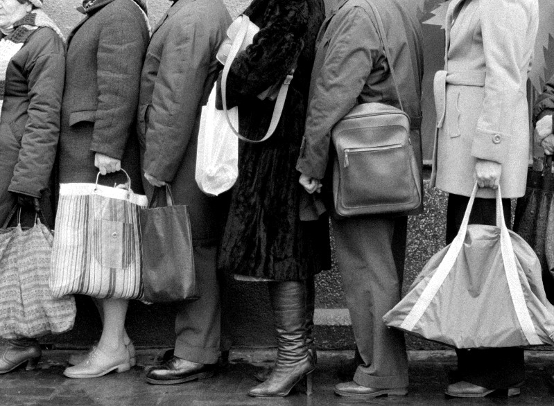 Czarno-białe zdjęcie przedstawia kilka osób stojących blisko siebie w kolejce, niosących duże torby i ubranych w płaszcze. Widoczne są tylko ich dolne partie ciała, co podkreśla ich nogi, torby i odzież wierzchnią.