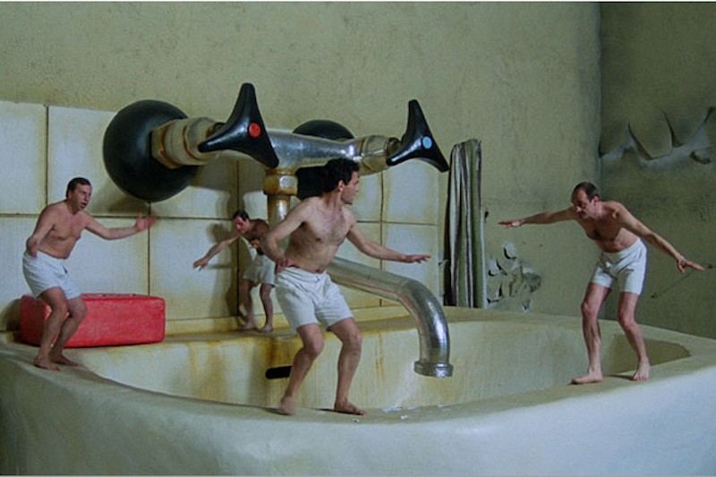 Czterech mężczyzn w białych spodenkach stoi na krawędzi gigantycznej umywalki, udając, że surfują. Za nimi górują ogromne umywalki, podkreślając surrealistyczną, zabawną scenę.