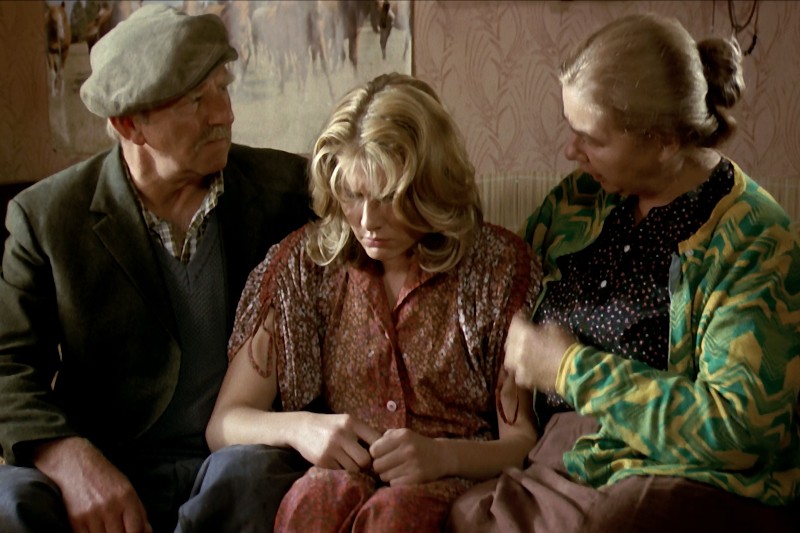 Młoda kobieta siedzi między starszym mężczyzną i kobietą na kanapie. Patrzy w dół, wyglądając na smutną, podczas gdy starsza para patrzy na nią z troską. Scena to skromny pokój z wzorzystą tapetą.