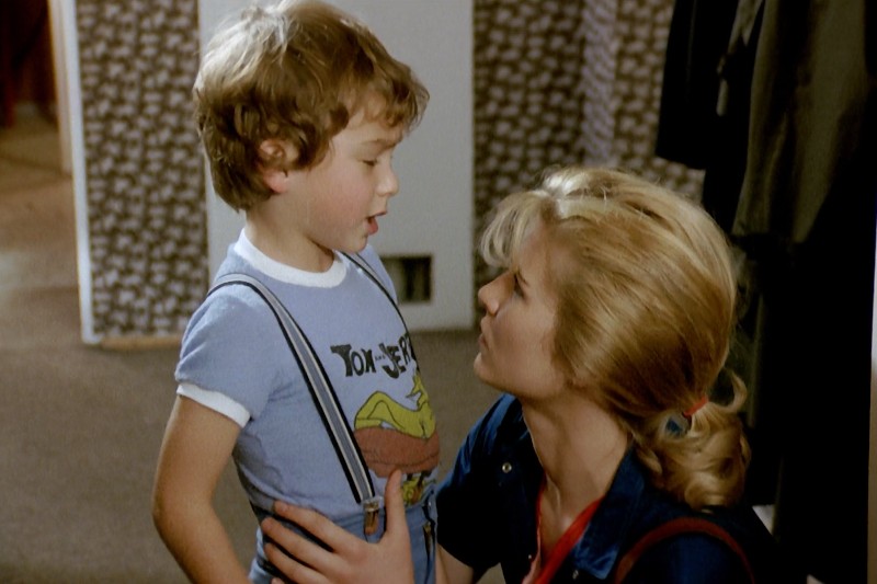 Kobieta z blond włosami spiętymi w kucyk klęka i patrzy na młodego chłopca ubranego w niebieski T-shirt z szelkami. Wygląda na to, że prowadzą szczerą rozmowę w pomieszczeniu.