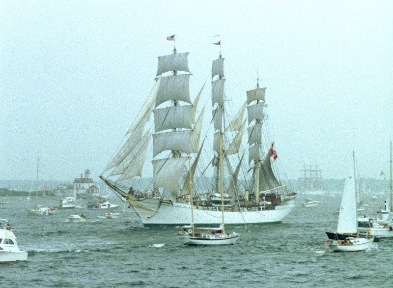 Duży, biały, wysoki statek z trzema masztami i pełnymi żaglami płynie po wodzie, otoczony kilkoma mniejszymi łodziami pod zachmurzonym niebem.
