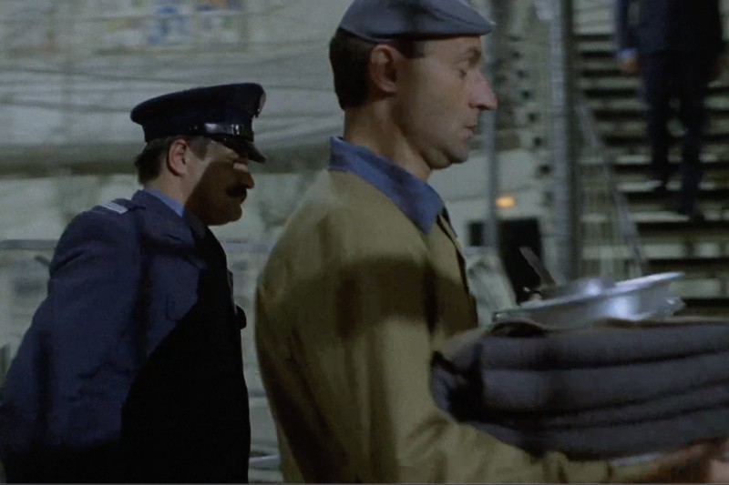 Mężczyzna w mundurze idzie za innym mężczyzną niosącym stos złożonych koców i tacek w pomieszczeniu, w tle widać schody.