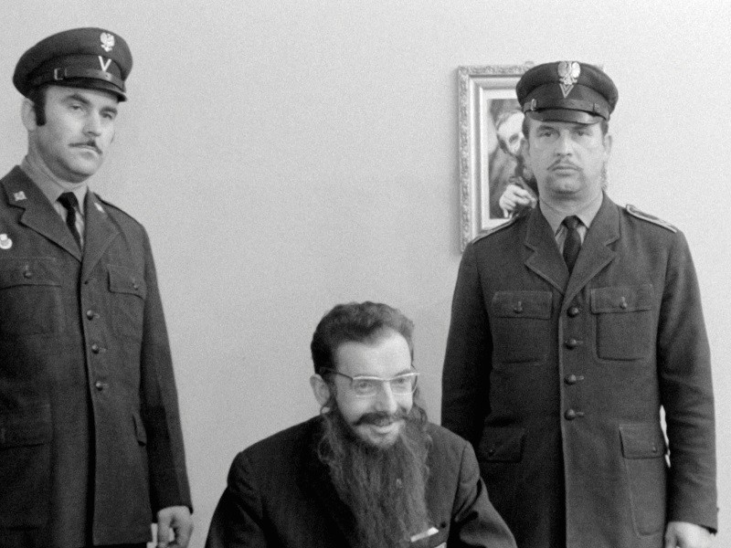 Trzech mężczyzn pozuje w pomieszczeniu. Dwóch stoi w mundurach z kapeluszami, a mężczyzna siedzący z przodu ma długą brodę i okulary. Na ścianie za nimi wisi oprawiony portret. Zdjęcie jest czarno-białe.