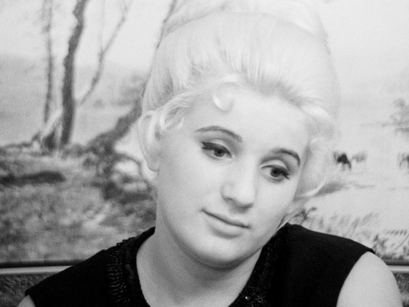 Kobieta z jasnymi włosami upiętymi w kok patrzy lekko w dół z łagodnym wyrazem twarzy. Ma na sobie ciemny top, a w tle widać rozmazaną scenę na zewnątrz.