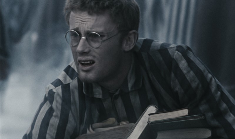 Zrozpaczony mężczyzna w okrągłych okularach i pasiastym więziennym uniformie ściska kilka książek, z zaniepokojonym wyrazem twarzy. Tło jest rozmazane i wydaje się ponure.