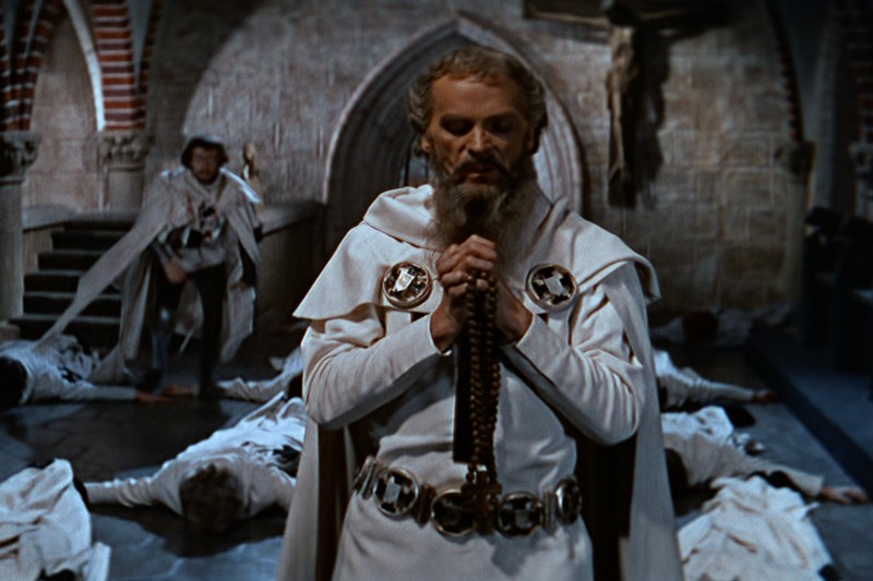 Brodaty mężczyzna w białej szacie z czarnymi krzyżami stoi i modli się, trzymając różaniec w kamiennej sali, podczas gdy inni w podobnych szatach leżą na ziemi za nim.