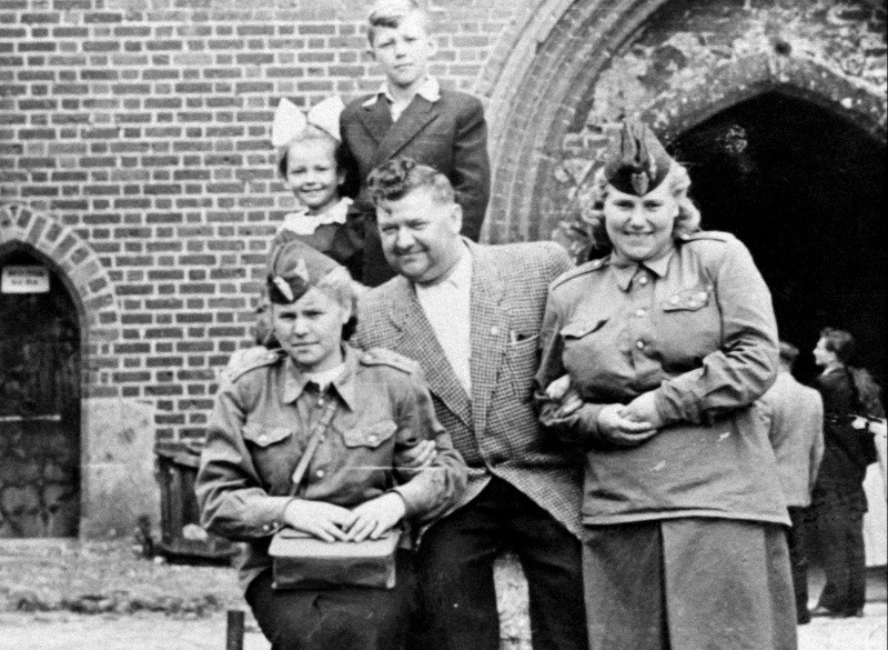 Czarno-białe zdjęcie przedstawia dwie kobiety w mundurach wojskowych, mężczyznę w kraciastej kurtce i dwójkę dzieci pozujących przed ceglanym budynkiem z łukowatymi drzwiami. Grupa stoi blisko siebie, uśmiechając się.