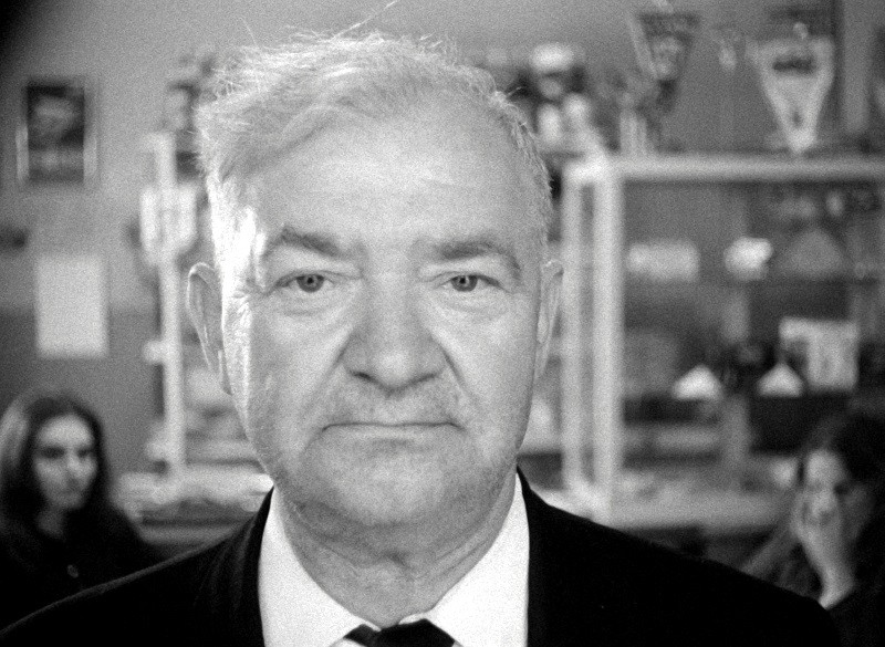 Czarno-białe zdjęcie starszego mężczyzny w garniturze i krawacie patrzącego prosto w kamerę. Tło jest lekko rozmyte, widać ludzi i półki z różnymi przedmiotami.