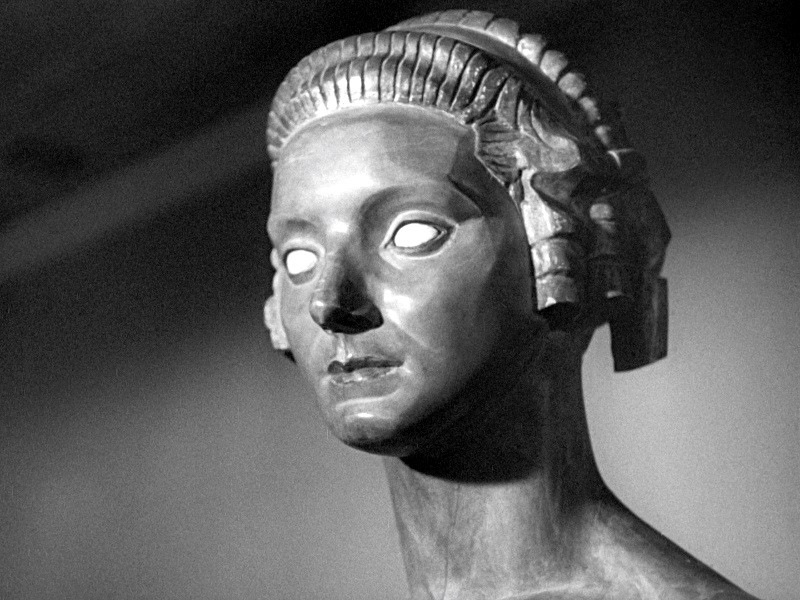 Zbliżenie kamiennego popiersia kobiety ze stylizowanymi włosami i pustymi, pozbawionymi źrenic oczami. Rzeźba ma gładkie rysy i pozbawioną wyrazu twarz, dramatycznie oświetloną w czerni i bieli.