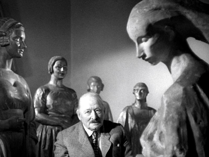 Starszy mężczyzna w garniturze siedzi otoczony kilkoma dużymi, ekspresyjnymi rzeźbami kobiecymi, patrząc lekko w górę i uśmiechając się. Scena jest dramatyczna z silnym oświetleniem i cieniami.
