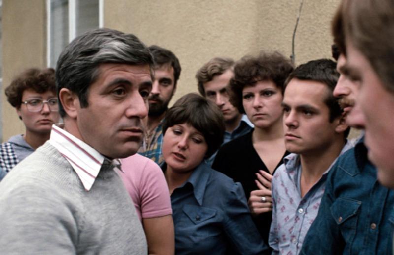 Mężczyzna w jasnym swetrze przemawia poważnie do grupy ludzi, którzy słuchają uważnie z zaniepokojonymi minami, stojąc blisko siebie na zewnątrz, niedaleko beżowego budynku.