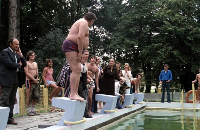 Grupa ludzi stoi i siedzi przy odkrytym basenie, obserwując mężczyznę w kąpielówkach przygotowującego się do skoku z bloku startowego. W tle widać drzewa.