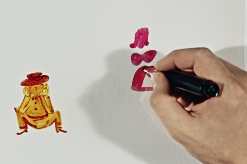 Ręka rysuje czerwonym markerem na białym papierze. Po lewej stronie znajduje się prosta, kreskówkowa postać żółtej żaby w brązowym kapeluszu. Po prawej stronie rysowane są różowe kształty.