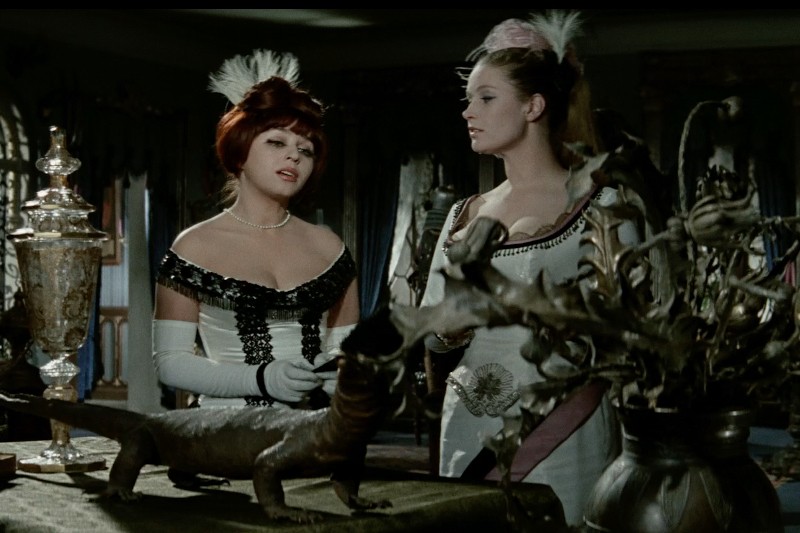 Dwie kobiety w wyszukanych, historycznych strojach stoją przy stole z ozdobnymi dekoracjami i małą rzeźbą smoka. Jedna kobieta ma na sobie czarno-białą suknię, druga białą i lawendową, obie noszą pióra na głowach.