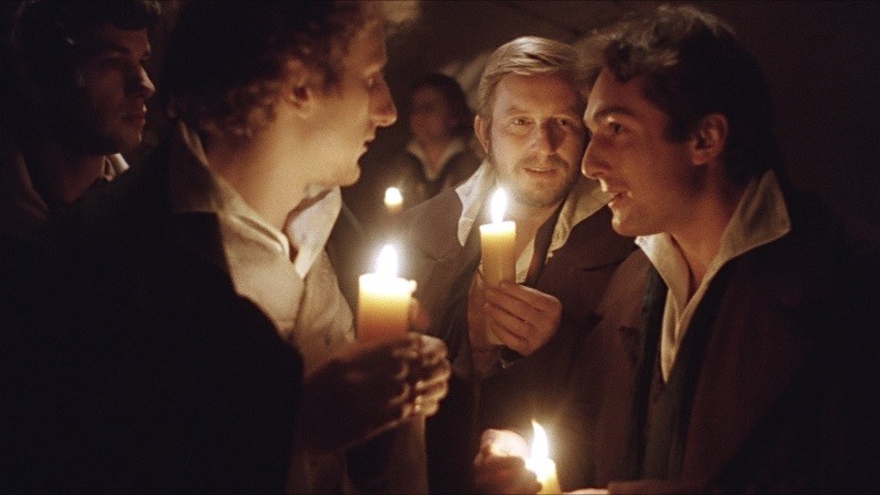 Czterech mężczyzn ubranych w stroje z epoki trzyma zapalone świece w słabo oświetlonym pomieszczeniu, rozmawiając. Ich twarze są delikatnie oświetlone światłem świec, co tworzy ciepłą i intymną atmosferę.