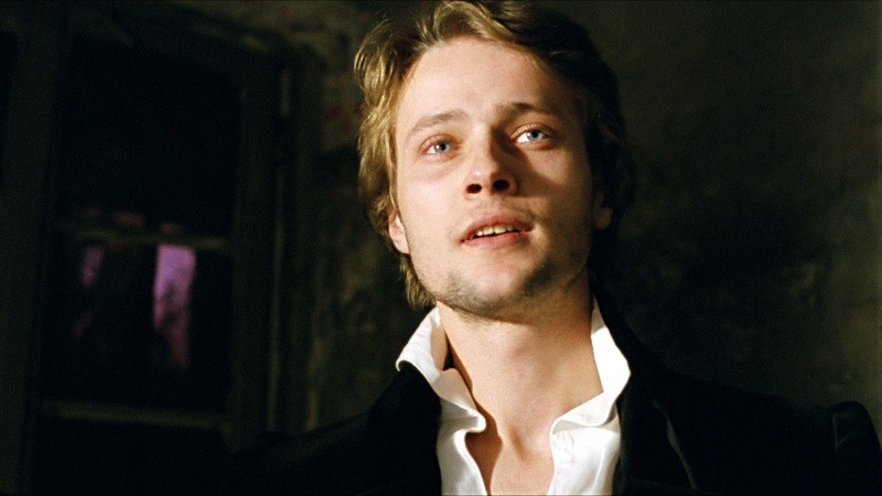 Młody mężczyzna o jasnych włosach i niebieskich oczach, ubrany w białą koszulę z rozpiętym kołnierzykiem i czarną kurtkę, stoi wewnątrz budynku, a jego twarz na ciemnym tle oświetla słabe światło.