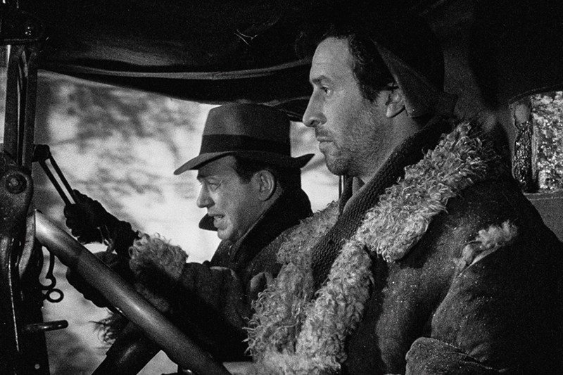 Dwóch mężczyzn siedzi na przednich siedzeniach pojazdu, obaj ubrani w grube płaszcze; mężczyzna po lewej ma na głowie kapelusz i prowadzi, podczas gdy mężczyzna po prawej patrzy przed siebie poważnie. Obraz jest czarno-biały.