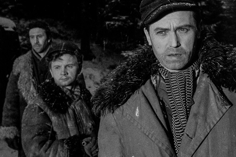 Trzech mężczyzn w grubych zimowych płaszczach i czapkach stoi na zewnątrz w zaśnieżonym, zalesionym terenie, wyglądając poważnie i skupieni. Czarno-biały obraz sugeruje napięty lub dramatyczny moment.