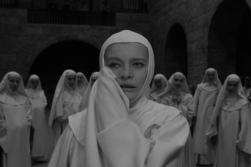 Grupa zakonnic w białych habitach stoi na kamiennym dziedzińcu. Centralna zakonnica wygląda na zmartwioną, trzyma rękaw przy twarzy, podczas gdy inne zakonnice stoją uroczyście w tle.