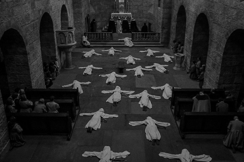 Grupa ludzi w białych szatach leży twarzą w dół z wyciągniętymi rękami na podłodze kamiennego kościoła, podczas gdy inni siedzą w ławkach, a trzy postacie stoją przy ołtarzu z przodu. Scena jest czarno-biała.