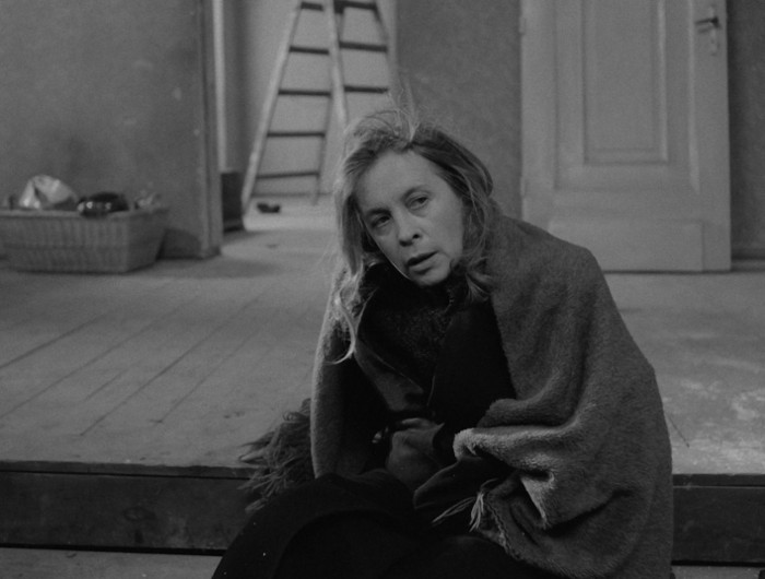 Kobieta z rozczochranymi włosami siedzi na podłodze owinięta w koc, wygląda na zmęczoną i przygnębioną, w skromnie umeblowanym, słabo oświetlonym pokoju z zamkniętymi drzwiami i drabiną w tle.