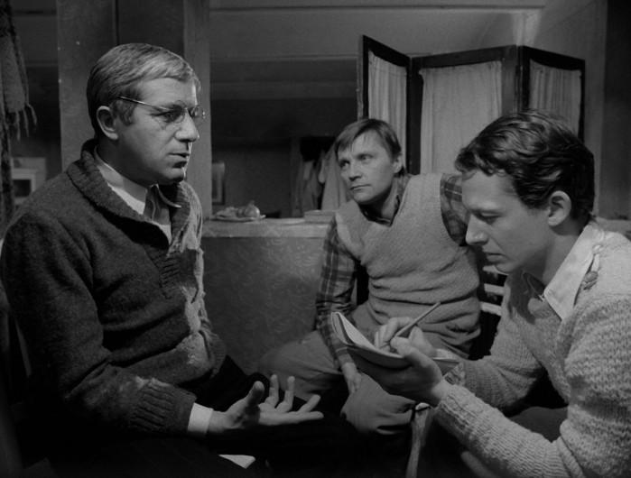 Trzech mężczyzn siedzi w pomieszczeniu na czarno-białej scenie; mężczyzna po lewej gestykuluje i rozmawia, mężczyzna w środku uważnie się przygląda, a mężczyzna po prawej coś pisze w notatniku.