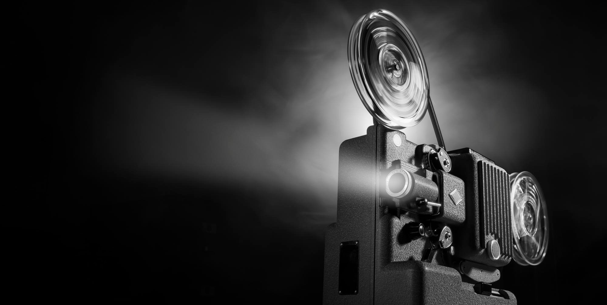 Zabytkowy projektor filmowy w czerni i bieli wyświetla światło i film, tworząc wiązkę, która świeci przez zadymione powietrze. Bębny obracają się podczas pracy projektora, przywołując klasyczną atmosferę kina.