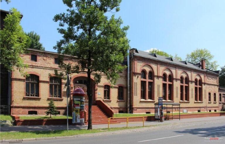 Długi, czerwony ceglany budynek z wysokimi łukowatymi oknami stoi przy ulicy otoczonej drzewami. Czerwone schody prowadzą do wejścia, a przystanek autobusowy z wiatą jest widoczny z przodu. Plakaty pokrywają pobliski cylindryczny kiosk.