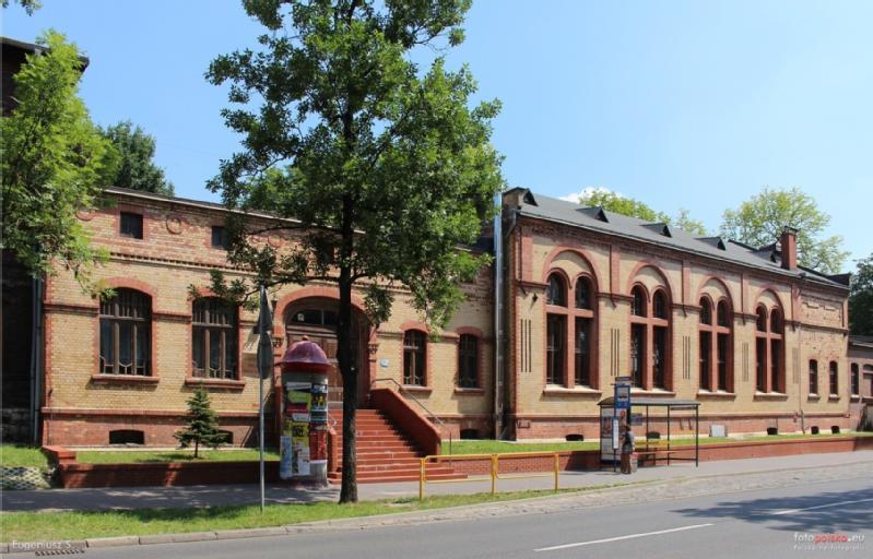 Długi, czerwony ceglany budynek z wysokimi łukowatymi oknami stoi przy ulicy otoczonej drzewami. Czerwone schody prowadzą do wejścia, a przystanek autobusowy z wiatą jest widoczny z przodu. Plakaty pokrywają pobliski cylindryczny kiosk.