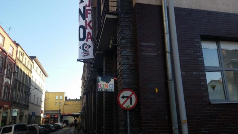 Widok ulicy pokazuje ceglany budynek z napisami KINO ZA ROGIEM i JFK. Na budynku zamontowany jest znak zakazu skrętu w lewo. Wzdłuż ulicy widoczne są inne budynki i samochody.
